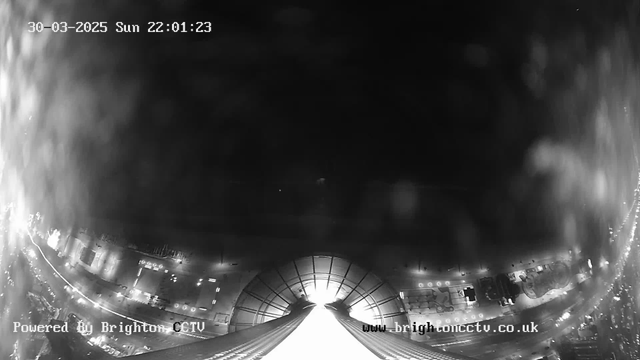 A black and white webcam image showing a low-angle view of a cityscape at night. The top part of the image is mostly dark, with some indistinct light sources in the background. The lower portion features a wide, illuminated area with diagonal lines leading towards a bright source at the center. The scene appears hazy, likely due to lens interference. There are faint outlines of buildings and structures visible along the edges. A timestamp in the corner indicates the date and time: March 30, 2025, at 22:01:23. The bottom of the image includes a watermark with "Powered By Brighton CCTV" and the website address.