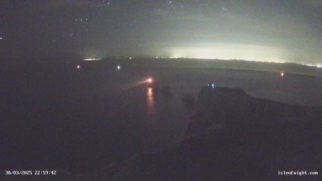 A nighttime seaside scene with a dark ocean and distant lights illuminating the horizon. Two small boats are visible, one with a bright orange light and another emitting blue light. The foreground features a rocky coastline silhouette against the shimmering water, reflecting the distant lights. The sky is filled with stars, though obscured by a slight haze.