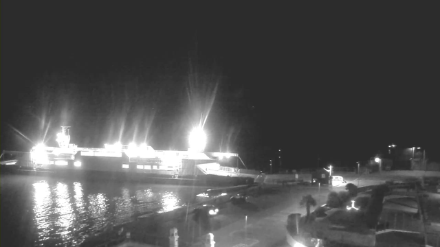A dark scene depicting a ferry docked at a shore. The ferry is brightly lit, with several lights illuminating its structure and the surrounding water. Reflected light shimmers on the surface of the water. The shoreline includes a few parked vehicles and structures in the background, partially lit by streetlights. The overall atmosphere is quiet, suggesting nighttime with no visible people around.