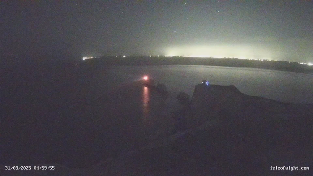 A dimly lit coastal scene at dawn, featuring a body of water with a visible boat emitting a red light. In the background, faint lights from a shoreline are visible, indicating a distant town. The sky is dark with scattered stars. The foreground shows a rocky cliff or landmass leading into the water. The timestamp at the bottom indicates the image was captured on March 31, 2025, at 4:59 AM.