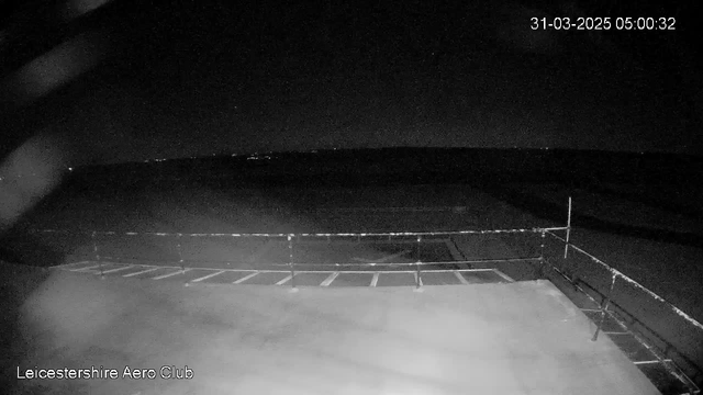 A dark scene captured by a webcam, showing a flat surface resembling a runway or platform. There is a railing along the edge, and beyond it, the night sky is visible with faint distant lights. The date and time displayed in the corner indicate it is early morning on March 31, 2025. The overall atmosphere is quiet and still, typical of a nighttime setting.