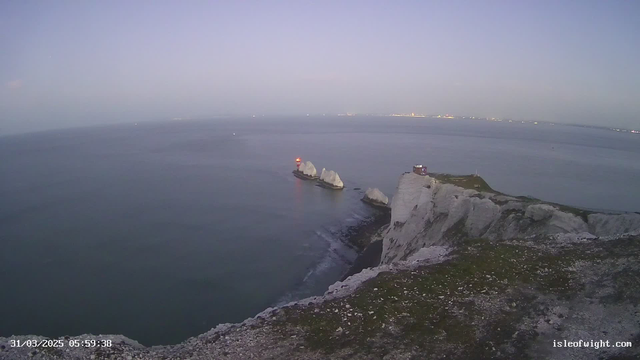 A scenic view of the coastline at dawn, featuring white chalk cliffs on the right side. In the foreground, several jagged rock formations jut out into the calm sea, where boats are visible in the distance. A red and white lighthouse stands on one of the rocks, with a small, wooden building on top of a nearby cliff. The sky displays soft hues of blue and purple as night transitions to day.