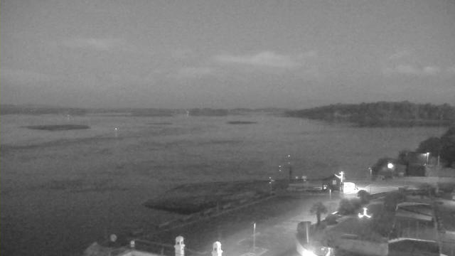 A black and white image captures a calm body of water under a cloudy sky, with distant land visible on the horizon. The foreground shows a harbor area with boats and a few buildings, illuminated by artificial lights. The overall scene is serene, with a hint of fog or mist over the water.