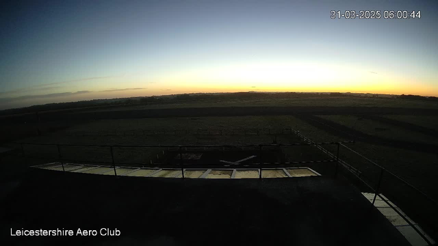A webcam view of a landscape at dawn, with a clear sky transitioning from dark blue at the top to lighter shades near the horizon. The sun is just below the horizon, casting a soft glow. In the foreground, there is a railing and a flat surface, possibly a viewing platform of the Leicestershire Aero Club. The ground is visible with some dark patches, likely indicating grass or fields, and a hint of a runway is seen in the distance.