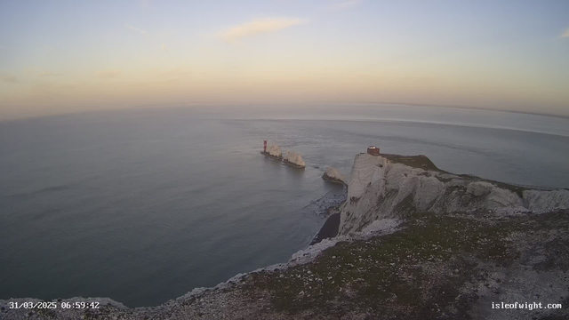 A tranquil seascape shows the calm ocean under a pastel sky, transitioning from light blue to soft yellow as the sun rises. In the foreground, a steep white cliff with grassy patches leads down to the water. Offshore, two distinctive rock formations, known as the Needles, display sharp edges and are topped with a red lighthouse on one side. The scene conveys a sense of peace and natural beauty as the day begins.