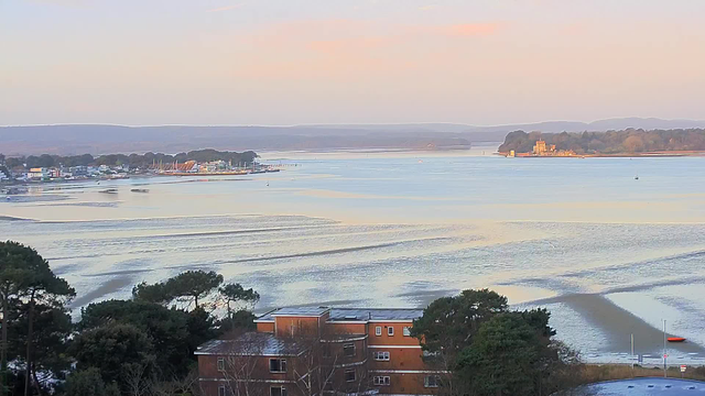 A serene landscape featuring a calm body of water that reflects soft hues of pink and orange from the sky. In the foreground, there are trees and a low building, while the shoreline includes a mix of small boats. In the distance, a small town can be seen with several buildings and masts of boats, leading to an area of land with trees and a structure that appears to be a castle or historic building. The scene conveys a peaceful, tranquil atmosphere.