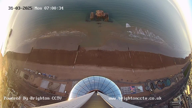 Aerial view of a beach during early morning. The scene captures calm, greenish waters with gentle waves lapping at the shore. In the center, a partially submerged wooden pier structure is visible. Below, the beach features a smooth, sandy area bordered by wooden posts. Along the bottom, there are hints of buildings and structures, including a circular design on the ground. The sky is brightening, indicating sunrise. The timestamp shows it is March 31, 2025, at 7:00 AM.
