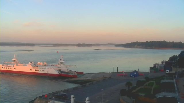 A serene waterfront scene at dawn, featuring a large white ferry with red accents docked at a pier. The calm water reflects soft hues of blue and pink from the morning sky. Small green islands are visible in the distance, and a few boats can be seen navigating the water. The right side of the image shows a coastal path lined with palm trees and other greenery, leading to a building near the edge of the pier.