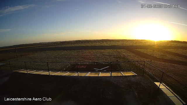 A wide view of a runway area at dawn, with the sun rising on the horizon, casting a warm orange glow. In the foreground, there is a railing and a paved area leading to a set of stairs. The ground is covered in dew, and there are grassy patches surrounding the runway, which is mostly clear. A small red windsock is visible on the left side of the image, with a smooth, flat strip of earth extending to the right. The sky is mostly clear with some wispy clouds.