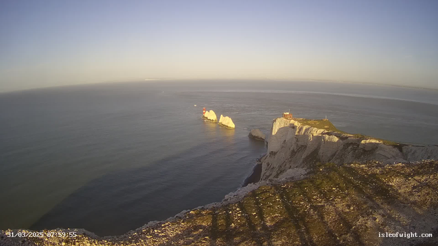 A scenic view of cliffs bordering a calm sea under a clear sky. The coastline features prominent white chalk cliffs with a lighthouse and a small red structure on the land. In the distance, a few large rock formations are partially submerged in the water. The light is soft, suggesting either early morning or late afternoon. Shadows from the cliffs stretch across the foreground, indicating the position of the sun.