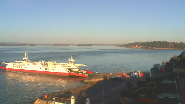 A ferry with a white and red exterior is docked at a pier, surrounded by calm water. In the background, there are hills partially obscured by mist, with a clear blue sky above. The foreground features a paved area with cars parked and small structures, suggesting a harbor setting. The scene conveys a tranquil morning atmosphere by the water.