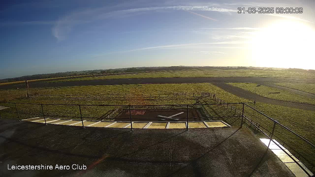 A wide view of an open field with a runway and grassy areas under a clear blue sky. The sun is rising on the right side, casting light over the landscape. In the foreground, there is a paved area with a helipad marked by a white cross. A fence runs along the edge of the field, and a small building is visible on the left side of the image. The timestamp in the top right corner shows the date and time.