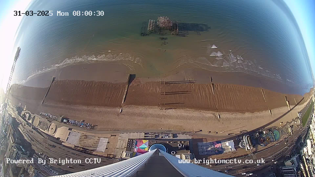 A webcam view captures the beach and shoreline at dawn. Below, there is a broad expanse of sandy beach with various structures, including wooden beams extending into the water. Gentle waves lap against the shore. In the distance, the ocean extends toward the horizon. The background shows the amusement park area with rides and colorful attractions along the promenade, and the image is marked with the date and time in the upper left corner. The scene is bright and clear, with a few scattered clouds in the sky.
