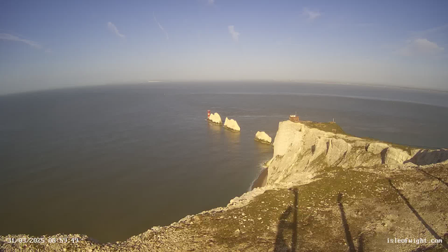 A coastal view featuring high white chalk cliffs on the right side of the image. Below the cliffs, the surface of the ocean is calm and reflects the blue sky. Two distinct rocks emerge from the ocean, one of which has a red lighthouse on it. In the background, the horizon stretches into the distance, with a few clouds scattered in the sky. The scene is serene, with no visible boats or human activity.