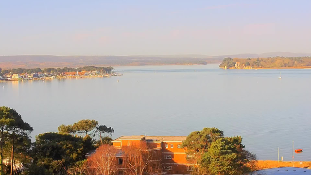 A serene view of a calm lake with gentle ripples reflecting the sky. In the foreground, there are buildings with red roofs surrounded by trees. Along the shoreline, a small marina is visible with several boats docked. The background features rolling hills and a clear sky, suggesting a peaceful early morning or late afternoon atmosphere.