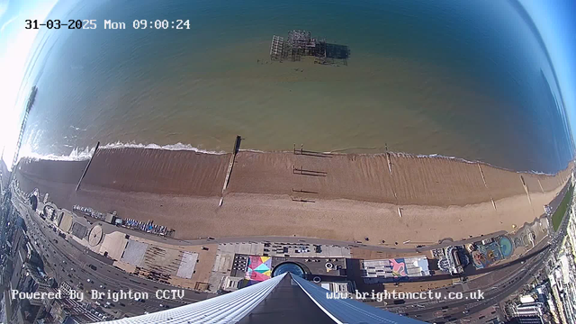 Aerial view of a sandy beach with a calm ocean. There are several wooden piers extending into the water, with one partially submerged in the distance. The beach is lined with beach huts and a promenade that includes various recreational areas and attractions. Above the beach, the sky is clear, and the time is displayed as 09:00 on March 31, 2025. The image has a digital watermark indicating it is powered by Brighton CCTV.