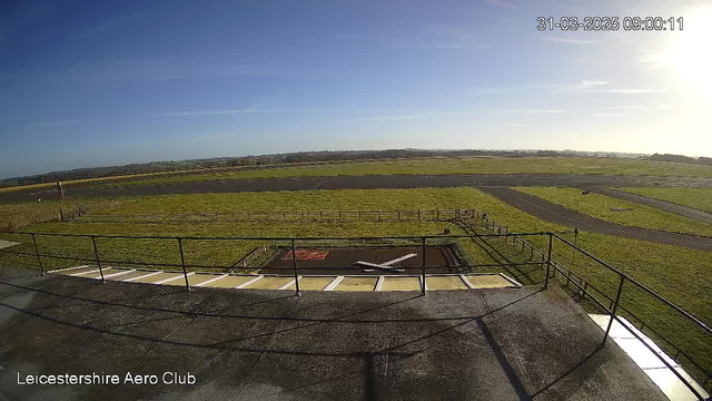 A wide view of a grassy airfield under a clear blue sky with faint wisps of clouds. In the foreground, there is a railing and a helicopter landing pad marked with an 'X'. The airfield stretches out in the background and is bordered by a fence, with a narrow road visible. The sun is shining brightly, creating a warm ambiance in the scene.