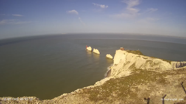 A scenic view of a coastline featuring prominent white chalk cliffs and the ocean. In the foreground, there is a grassy area at the cliff's edge. To the right, a small lighthouse stands on a rocky outcrop near the water, with its red and white striped design visible. The sky above is bright blue with a few wispy clouds. The water appears calm, reflecting some light, and the horizon stretches into the distance. In the background, there are distant land masses faintly visible on the horizon.