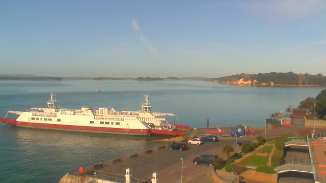 A ferry with a white hull and red trim is docked on calm water. The background features a shoreline with wooded hills and a cliff visible in the distance. In the foreground, vehicles are parked on a paved area, and there are palm trees and grassy areas. The sky is clear and blue with a few wispy clouds.