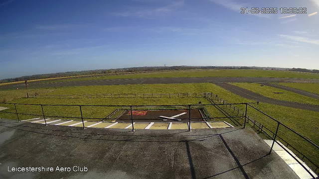 A wide view of an airport runway surrounded by green grass under a clear blue sky. In the foreground, there is a railing and a paved area with a white X marking a landing spot. The runway extends into the distance, with a few small structures and a wind direction indicator visible on the right side.