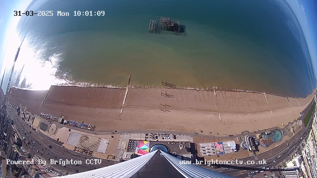 A high-angle view shows a beach with a long stretch of sand and a gentle sea wave lapping at the shore. There are several people walking along the beach. Offshore, a partially submerged structure is visible in the water. The scene includes a colorful area with tents and structures near the beach, alongside a road lined with vehicles. The sky is clear with some blue and slight clouds. A timestamp in the upper left corner indicates the date and time.