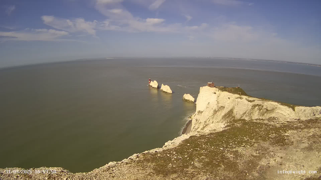 A scenic view of a coastal landscape featuring white chalk cliffs. In the foreground, the cliffs rise sharply with some grassy areas. Off the coast, there are two large rocky formations, one of which has a red and white lighthouse on it. The sea is calm and reflects the sky above, which is mostly clear with a few scattered clouds. The horizon is visible in the distance, where the water meets the sky.