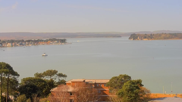 A calm water scene featuring a wide, tranquil bay surrounded by rolling hills. In the foreground, there are scattered trees and a low, brick building with a flat roof. Boats are visible on the water, with one sailboat prominently anchored in the middle distance. To the left, a marina filled with small boats and colorful homes lines the shoreline. In the background, an island with trees and a structure can be seen, along with more distant hills under a clear blue sky.