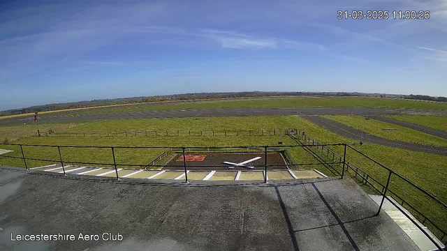 A view from a webcam at the Leicestershire Aero Club, showing a wide expanse of green grass and a clear blue sky. In the foreground, there is a fenced area with a white cross-shaped object, possibly representing a runway marker. A section of the tarmac airstrip is visible, leading into the distance where more grassy areas and a distant tree line can be seen. The time and date are displayed in the top right corner, indicating it is 11:00:26 on March 31, 2025.