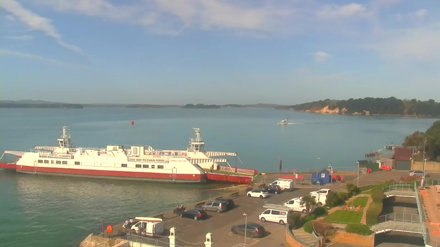 A scenic waterfront view featuring a large ferry docked at a pier. The ferry is primarily white with red and grey accents, displaying signage indicating speed and navigational instructions. In the background, a calm blue waterway extends towards distant green hills under a clear sky with a few wispy clouds. On the mainland, there are several parked vehicles, including cars and a van, along with a well-maintained path and grassy areas near the waterfront. A small boat is visible in the water, moving away from the ferry.
