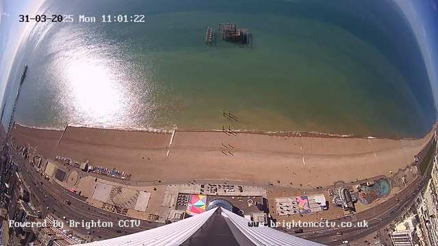 A wide-angle view from a high vantage point overlooking a beach with golden sand and turquoise water. The beach is populated with small figures walking along the shoreline. A pier extends into the water on the left side of the image. In the distance, there is a partly submerged structure in the water. The area below the camera shows colorful buildings and attractions along the beachfront, with a geometric pattern on the ground. The sky is mostly clear with a few wispy clouds. The date and time are stamped at the top of the image.