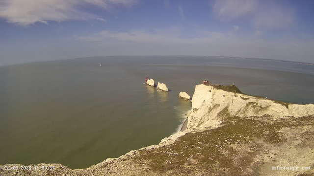 A scenic view of the coast features white chalk cliffs meeting a calm sea under a bright blue sky with scattered clouds. In the distance, there are two prominent white rock formations with a small building and a red lighthouse on one. The foreground shows a rocky landscape with patches of grass. A small sailboat is visible on the water.