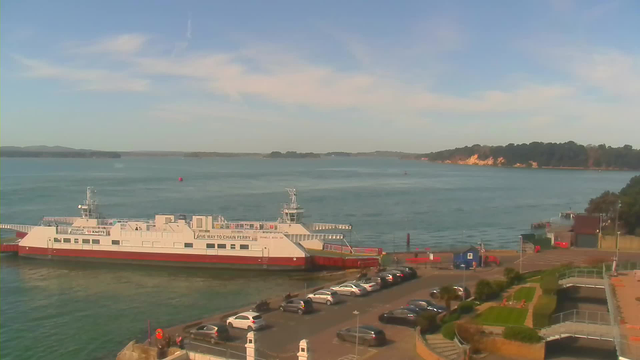 A ferry is docked at a harbor, with the body of water extending into the background. The ferry is white with red accents and has a sign that reads "SQUAD 2 CHAIN FERRY". There are a few people visible on the ferry. In the foreground, there is a parking area filled with cars. To the right, there are green grassy areas and landscaped paths leading toward the water. In the distance, hills and trees line the edge of the opposite shore under a clear blue sky.