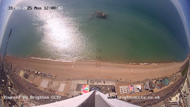 A high-angle view of Brighton's coastline on a sunny day. The scene features a sandy beach with scattered people walking along the shore. The blue-green water reflects sunlight, creating a shimmering effect. In the background, the remains of a pier can be seen in the water. The beach area includes various colorful structures and attractions, with a pathway running parallel to the beach. The image includes a timestamp and a watermark at the bottom.