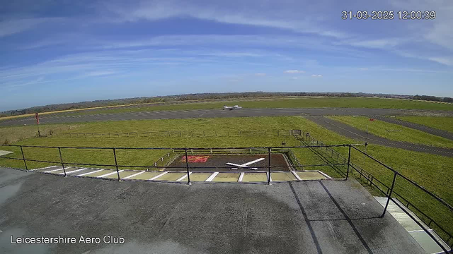 A clear blue sky with a few clouds above a grassy airfield. In the foreground, there is a railing along a rooftop, and below it, a marked spot with a large white "X" on the ground. An aircraft is taxiing on the runway, which is mostly paved and stretches into the distance. To the right, there is a windsock indicating wind direction. The landscape is open with sparse fencing and trees in the background. The image has a timestamp indicating the date and time.