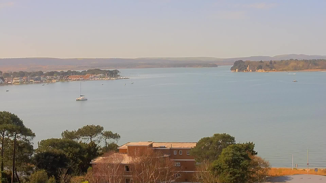 A calm body of water stretches across the image, with a gentle shoreline lined with trees and some buildings. Several boats are scattered on the water, including a small motorboat and a sailboat. In the background, a series of distant hills are visible beneath a clear sky. The scene conveys a peaceful waterfront view, illuminated by natural light.