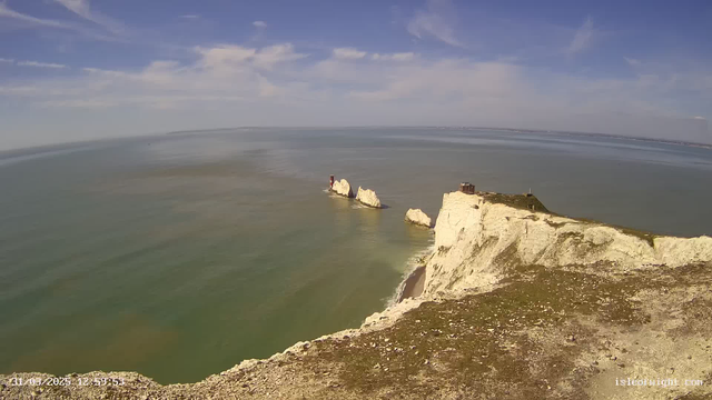 A coastal scene featuring white chalk cliffs rising from the blue ocean under a clear sky. In the background, distant land is visible on the horizon. Three small rocks protrude from the water, and a red structure is situated on one of the rocks. The foreground shows grassy land leading down to the coastline, which has a small sandy beach. The overall atmosphere is calm and serene.
