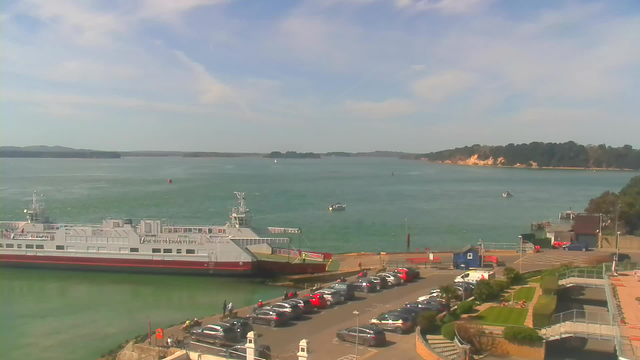 A serene waterfront scene featuring a large ferry docked at a pier, with several smaller boats visible in the water. In the foreground, a parking area is filled with vehicles, while pathways and grassy areas are seen nearby. The backdrop includes a smooth, blue sea with distant islands and a coastline of trees and cliffs under a clear sky with soft, light clouds.