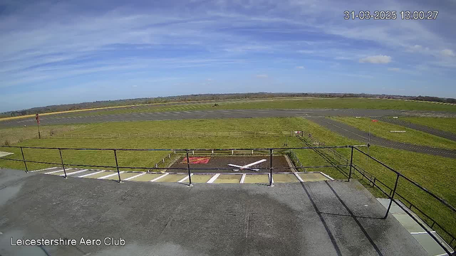 An open grassy area with a runway in the foreground and blue skies above. The runway has a dark pavement, with grassy patches around it. A white 'X' marking is visible on the ground, indicating a helipad or landing area. There is a low railing in the foreground. In the distance, there are some clouds and a gentle slope of land, contributing to the scenery. The image is bright and captures a clear day.