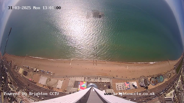 A high-angle view overlooking a sandy beach with a bright blue sea. The shoreline features gentle waves and several people walking along the beach. Below, there are colorful beach huts and a funfair with amusement rides. The image also includes a boardwalk with chairs and tables, as well as some visible buildings adjacent to the beach. The sky is relatively clear, indicating a sunny day. The time stamp in the top left corner reads "31-03-2025 Mon 13:01:00."