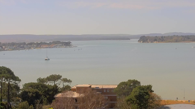 A coastal scene featuring a calm bay with a few boats anchored. In the foreground, there are trees and buildings, while the background showcases a distant shoreline with more boats and gently rolling hills. The sky is clear with a light gradient, indicating a sunny day.