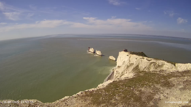 A panoramic view of a coastline featuring white chalk cliffs against a blue sky with scattered clouds. The water is calm and reflects shades of green and blue. In the distance, there are several rocky outcrops extending into the sea. On one of the outcrops, a small structure resembling a lighthouse is visible. The foreground consists of grassy terrain leading down to the beach.