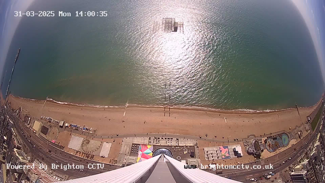 Aerial view of a beach with a wide, sandy shore extending along the bottom of the image. The water is a shimmering blue, reflecting sunlight, and a gentle wave pattern is visible near the shore. In the distance, a pier extends into the water, partially submerged and with some structure visible above the surface. There are people scattered along the beach and walking on a promenade. Various colorful beach huts and buildings line the beach, and a large, multicolored structure is also present on the sand. The sky is mostly clear with a few wispy clouds.