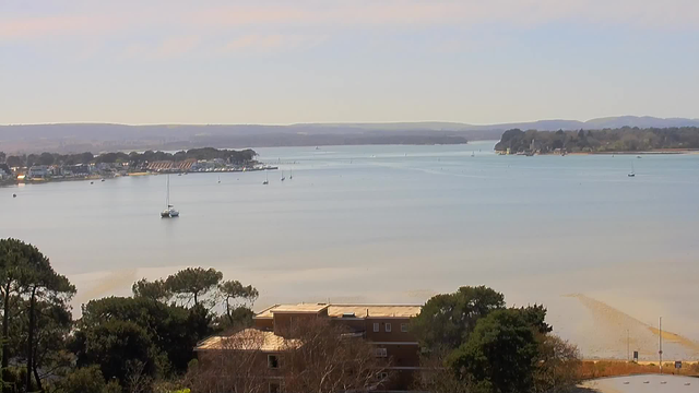 A panoramic view of a calm waterway, with sailboats scattered across the surface. In the background, a small town with houses and marinas lines the shore, surrounded by trees. Gentle hills rise in the distance under a clear sky with soft clouds. In the foreground, there are shrubs and trees near a building, partially obscured. The overall scene conveys a peaceful and natural atmosphere.