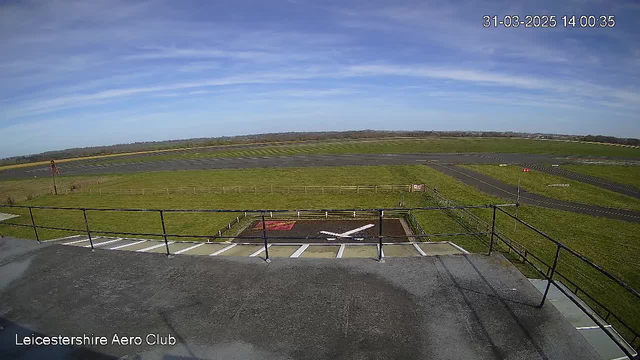 A view from a raised platform overlooking an airfield. The scene shows a clear blue sky with wispy clouds. Below, there is a grass-covered runway with taxiways, and a white X-shaped marking on the ground, likely indicating a landing area. Several flags are visible, with one red flag standing out. The area is bordered by a wooden fence, and there are no aircraft visible in the image. The time displayed is 14:00:35 on March 31, 2025.