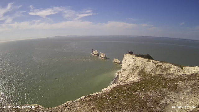 A panoramic view of a coastal landscape featuring white chalk cliffs. Below the cliffs, there is a sandy beach beside a calm turquoise sea with gentle waves reflecting sunlight. In the distance, a pair of prominent rock formations rise from the water, with one featuring a tall, red lighthouse. The sky is mostly clear with a few scattered clouds, providing a bright and sunny atmosphere. The scene captures the natural beauty of the coastline, with greenery on the cliff edges and a serene ocean horizon.