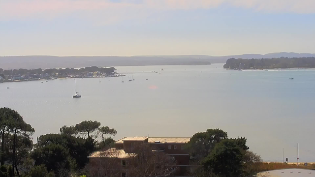 A serene view of a waterway with multiple small boats scattered across the calm surface. In the foreground, there are trees and rooftops partially visible, leading to a tranquil landscape that extends to distant hills under a clear blue sky. The sunlight creates a shimmering effect on the water, enhancing the peaceful atmosphere of the scene.