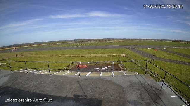 A wide view of an airport runway with green grass surrounding it under a clear blue sky. In the foreground, there is a helipad marked with a large white cross on a brown surface. A wooden railing borders the edge of the viewing area. The runway extends into the distance, with a few small aircraft seen far away on the left side of the image. The scene conveys a sense of openness and tranquility.