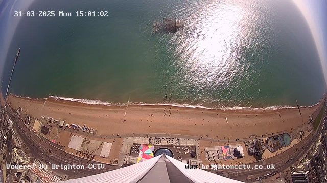 An aerial view of a sandy beach along the coastline, with gentle waves lapping at the shore. There are people enjoying the beach, visible as small figures scattered on the sand. To the left, a pier extends into the water, with some structures on it. On the shore, various colorful umbrellas and beach items are visible, along with rides and attractions nearby. The sky is clear with a few clouds, and the sunlight reflects off the water, creating a shimmering effect. The image is timestamped, indicating the date and time at the top.