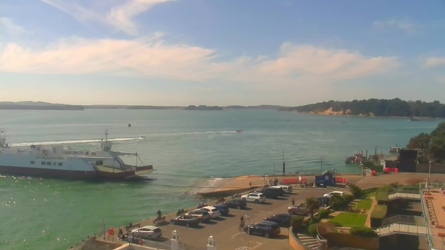 A view of a calm harbor with a large white and gray ferry docked on the left side. A small boat is creating a wake as it moves across the water. Cars are parked in a lot near the water, and a few people are walking along the shore. In the background, there are green hills and a clear blue sky with scattered clouds.