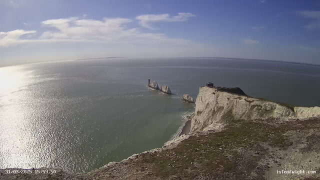 A panoramic view of a coastal scene featuring white chalk cliffs leading down to a calm sea. In the distance, there are several rock formations jutting out of the water. The sky is mostly clear with thin clouds, and the sunlight reflects on the ocean surface, creating sparkles. On the cliff's edge, a small building is visible, and the overall setting conveys a sense of tranquility and natural beauty.